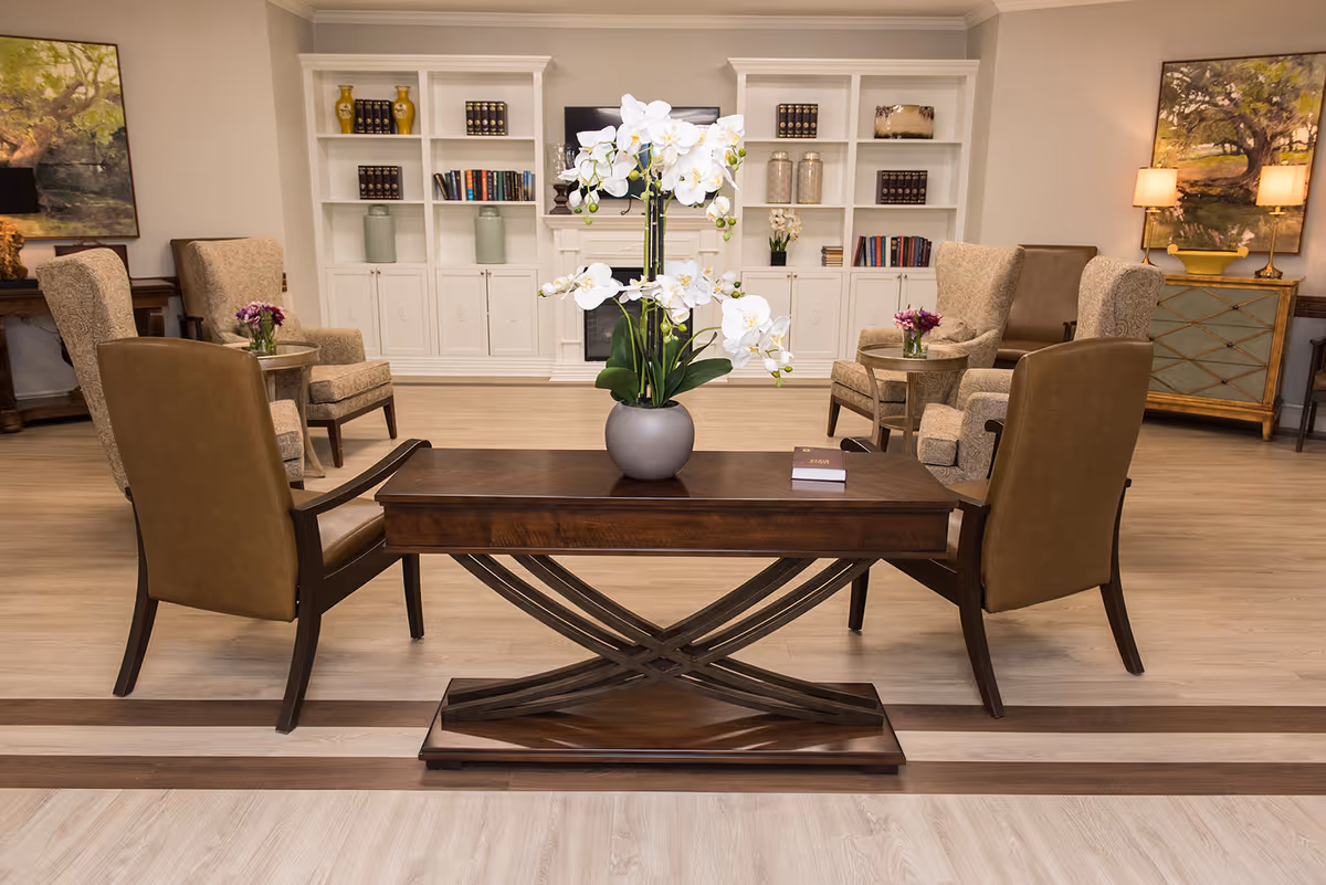 A well-furnished communal lounge with armchairs arranged around a central table topped with a vase of white orchids and built-in bookshelves in the background.