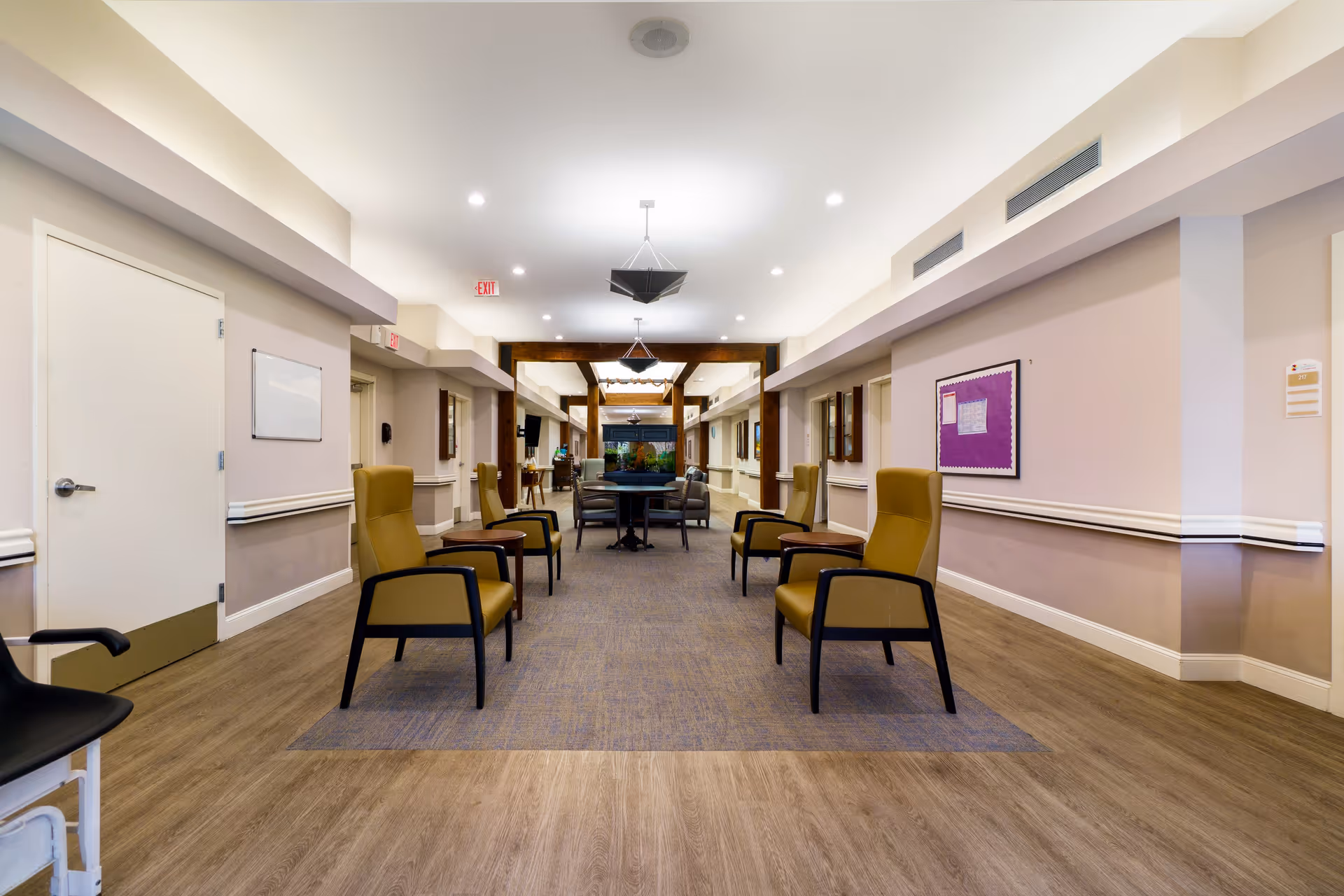 A spacious hallway in a senior living facility with several mustard-colored armchairs arranged along the sides. The floor is a combination of wood and carpet, and the walls are painted in light beige tones with white trim. The ceiling has recessed lighting and hanging light fixtures. There is a bulletin board on the right wall and a door on the left. At the far end of the hallway, there is a table with chairs and a large fish tank.