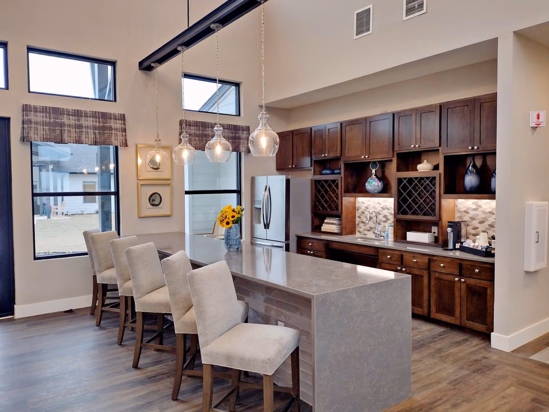 Modern kitchen area with a long gray marble island countertop surrounded by four beige upholstered chairs. Above the island hang four glass pendant lights. The back wall features dark wooden cabinets, a stainless steel refrigerator, a small sink, wine racks, decorative vases, and a coffee station. Large windows with patterned valances allow natural light into the space.