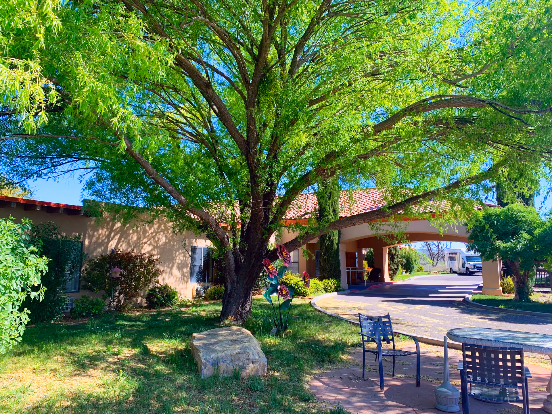 Outdoor view of Desert Peaks Assisted Living and Memory Care facility showing a large tree with green leaves, a stone bench, metal chairs and a table on a paved area, and the building entrance with a covered driveway and a vehicle parked in the background.