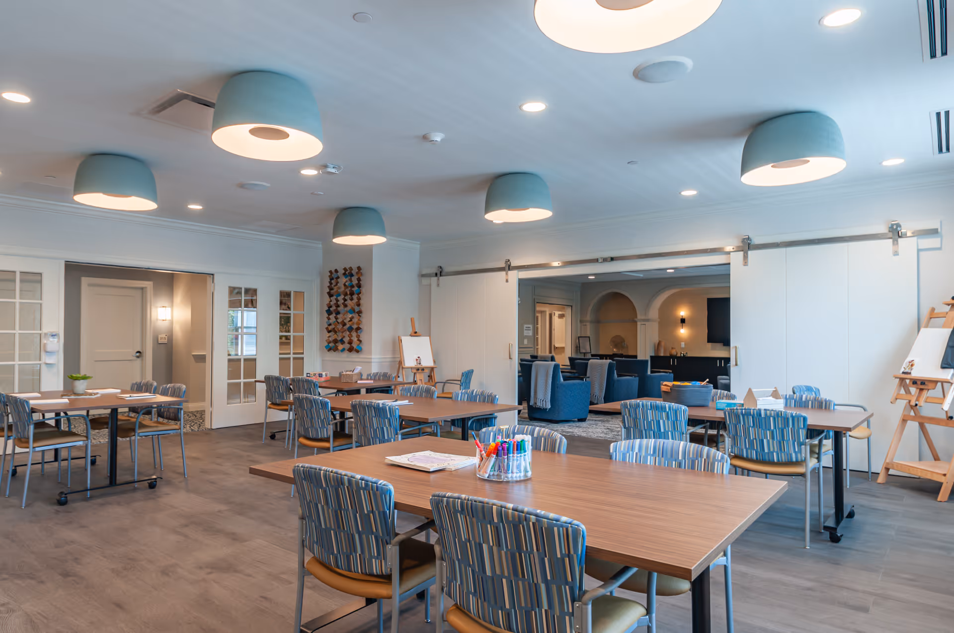 A bright and spacious activity room in a senior living facility with multiple tables and chairs arranged for group activities. The tables have art supplies like markers and paper. The room features modern ceiling lights, large sliding doors leading to a lounge area with comfortable blue armchairs, and an easel with a blank canvas. The flooring is wood-style, and the walls are painted light colors, creating a welcoming atmosphere.