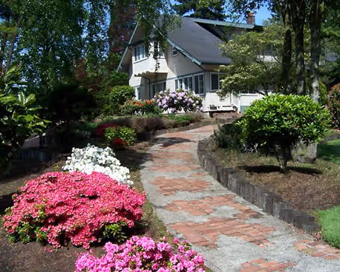 A charming house with a sloped roof surrounded by lush greenery and colorful flowering bushes along a brick and concrete pathway leading to the entrance.
