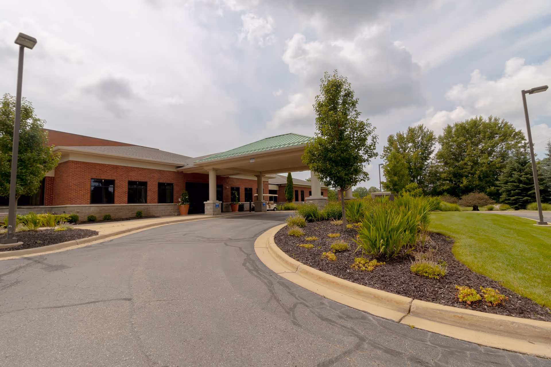 Covered entrance and circular driveway of a brick senior living facility with landscaped beds and trees.