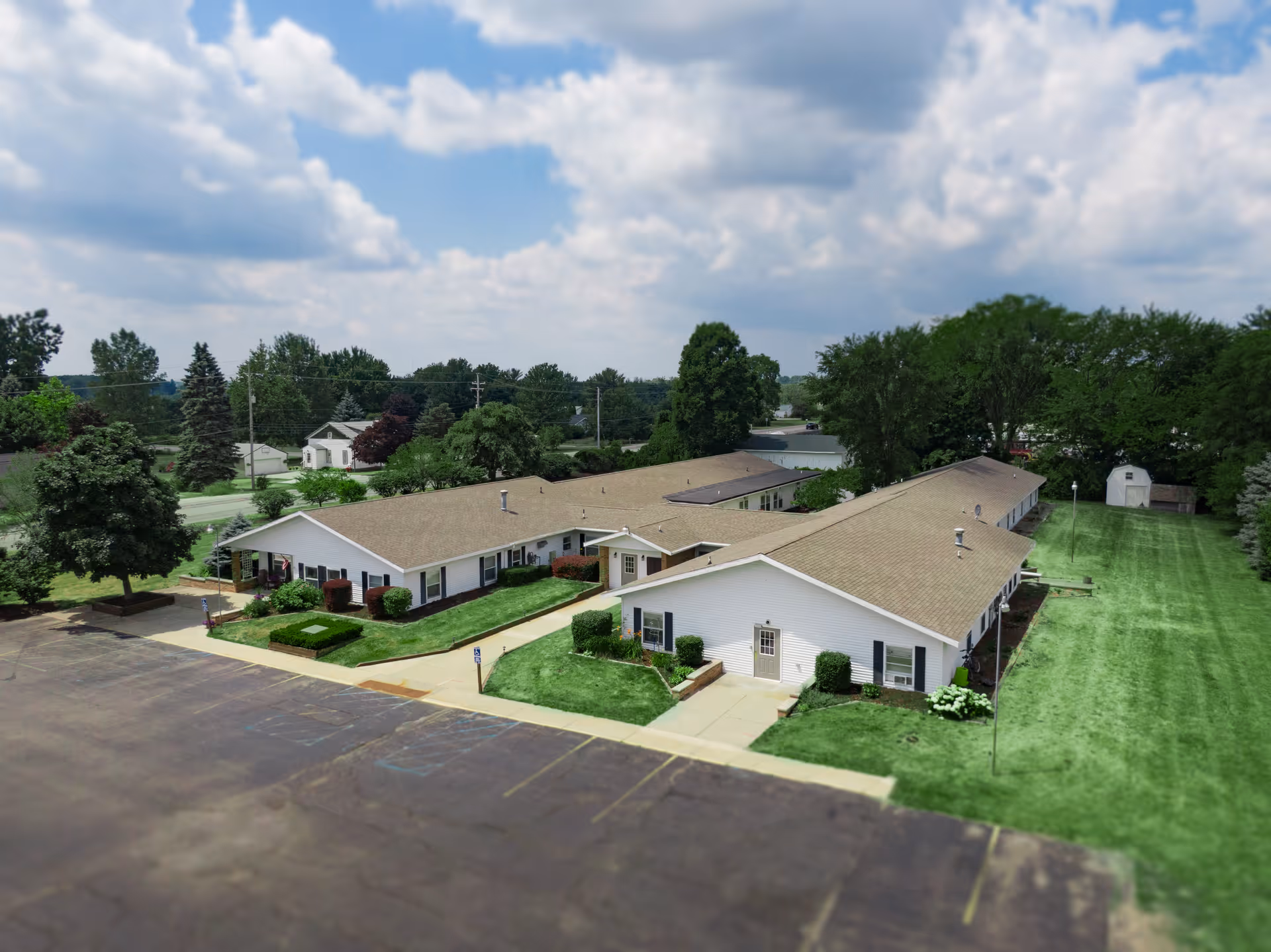 Aerial view of a single-story senior living facility with beige roofs and white exterior walls, surrounded by green lawns and trees under a partly cloudy sky. The building is shaped in an L-formation with paved walkways and a parking lot in the foreground.
