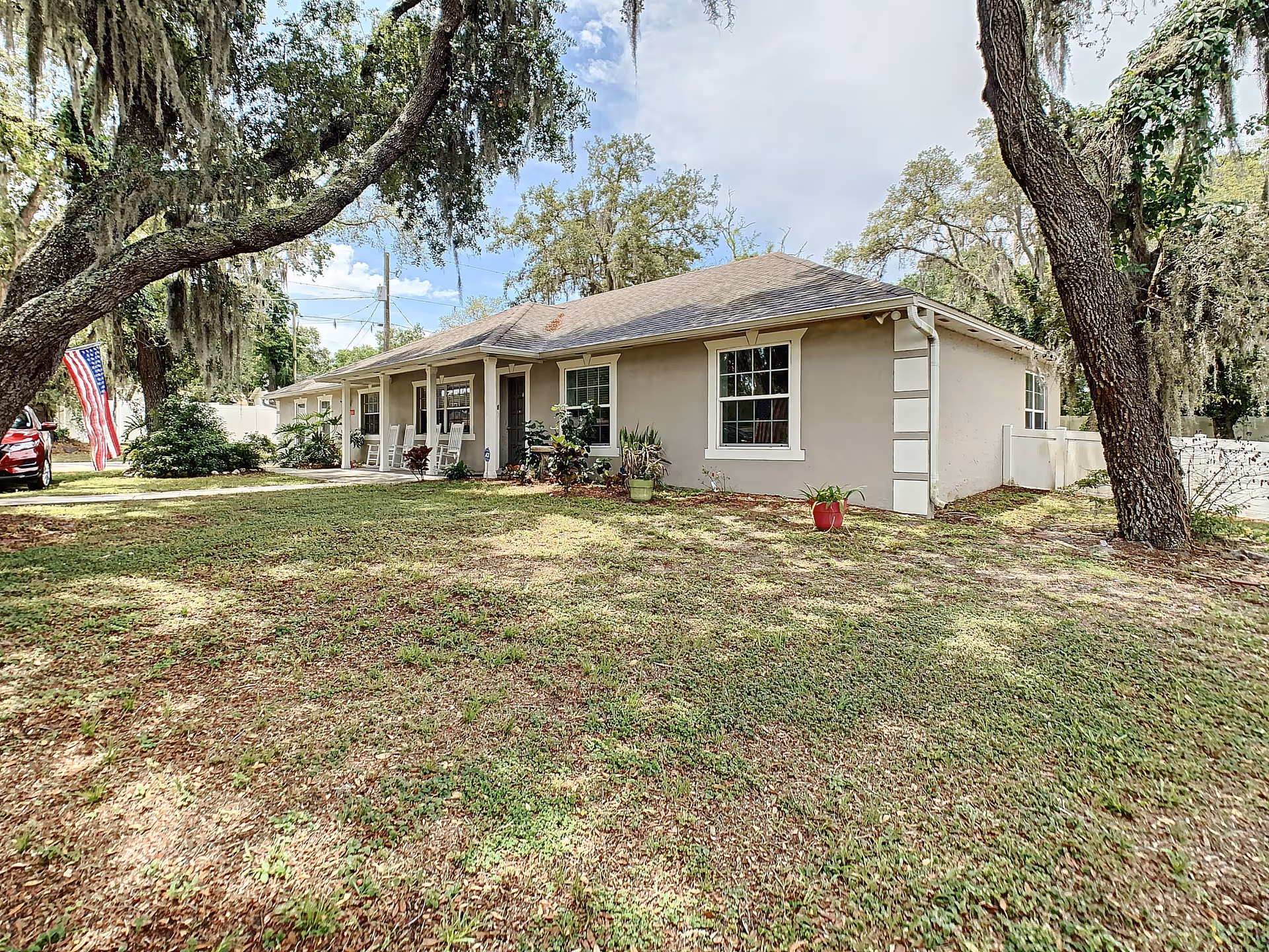 Single-story beige residential building with a front porch, lawn, large oak trees, and an American flag.