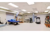A spacious rehabilitation room with various exercise and therapy equipment including a treadmill, stationary bike, therapy tables, and parallel bars. The room has white walls, a tiled ceiling with fluorescent lighting, and curtains that can be drawn around the therapy tables for privacy.