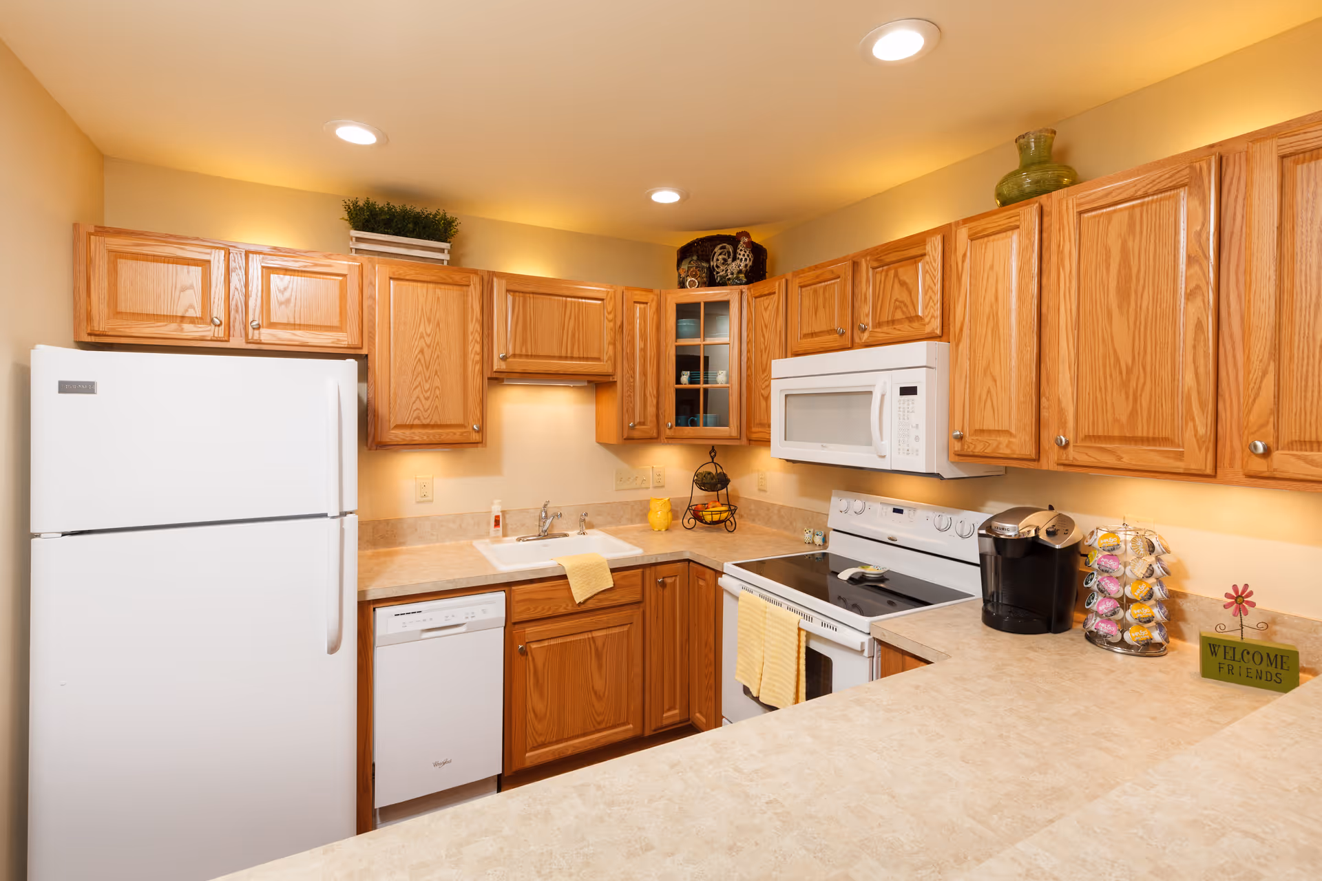 Well-lit kitchen with oak cabinets, white appliances, a countertop with a coffee maker and decorative items.