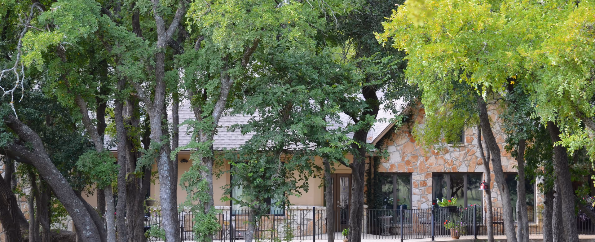 Exterior view of a stone and stucco building partially obscured by large green trees with a fenced patio area in front.