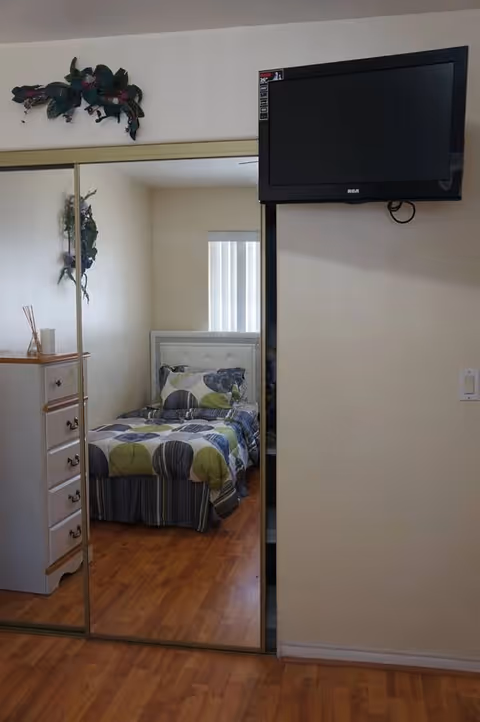 A small bedroom with a single bed covered in a patterned bedspread with green, gray, and white colors. There is a white dresser with multiple drawers on the left side, and a large mirrored sliding closet door reflecting the bed and part of the room. A flat-screen TV is mounted on the wall to the right above a light switch. The room has wooden flooring and light-colored walls.