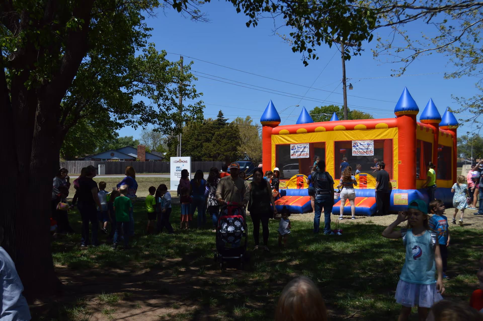 Outdoor scene at The Commons facility showing a group of people, including children and adults, gathered around a colorful inflatable bounce house on a sunny day with trees and a clear blue sky.