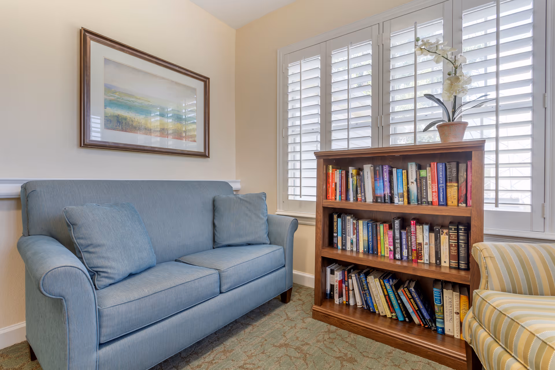 Light-filled sitting area with a blue sofa, wooden bookshelf full of books, and shuttered windows.