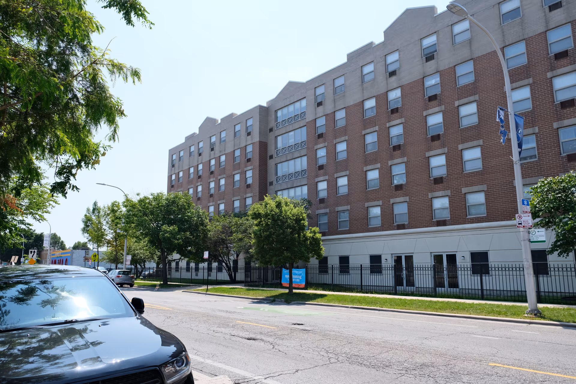 Exterior view of a multi-story brick and concrete building with numerous windows, trees lining the sidewalk, a black car parked on the street, and a clear blue sky.