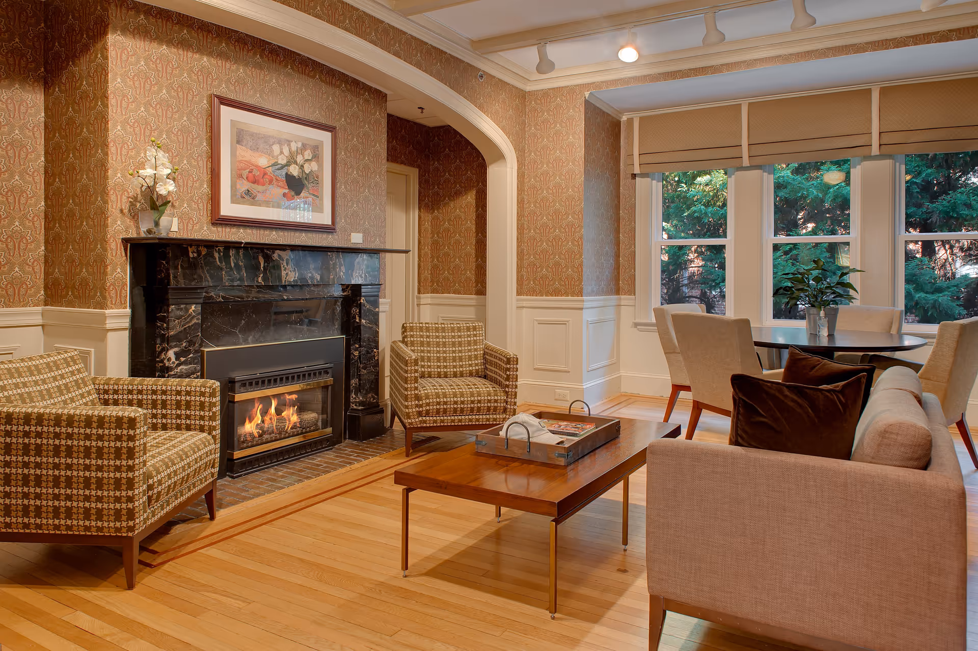 A cozy living room with a lit fireplace surrounded by a black marble mantel. Two patterned armchairs are positioned near the fireplace, and a beige sofa with brown pillows faces a wooden coffee table. Behind the sofa, there is a round dining table with four beige chairs, and large windows with beige blinds reveal green trees outside. The walls have a patterned wallpaper above white wainscoting, and a framed painting hangs above the fireplace.