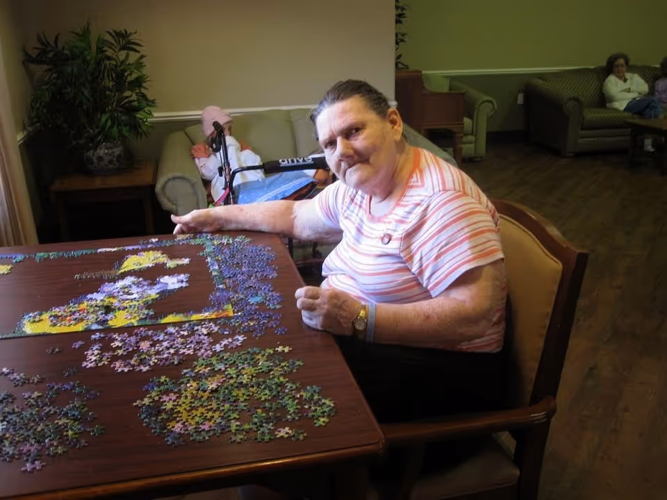 An elderly woman sitting at a wooden table working on a jigsaw puzzle in a common room with couches and other seated people in the background.
