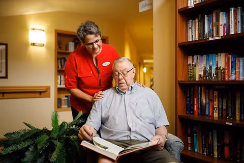 An elderly man sitting in a chair reading a book while a woman in a red shirt stands behind him with her hands on his shoulders, smiling. They are in a cozy room with bookshelves filled with books and warm lighting.