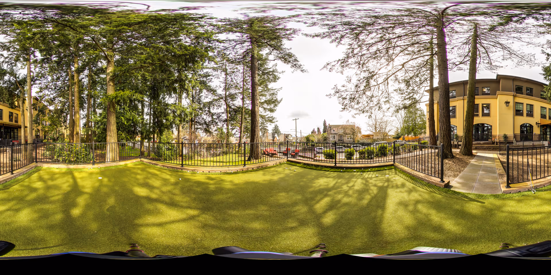 Outdoor fenced area with artificial grass surrounded by tall trees and a yellow building in the background under a partly cloudy sky.