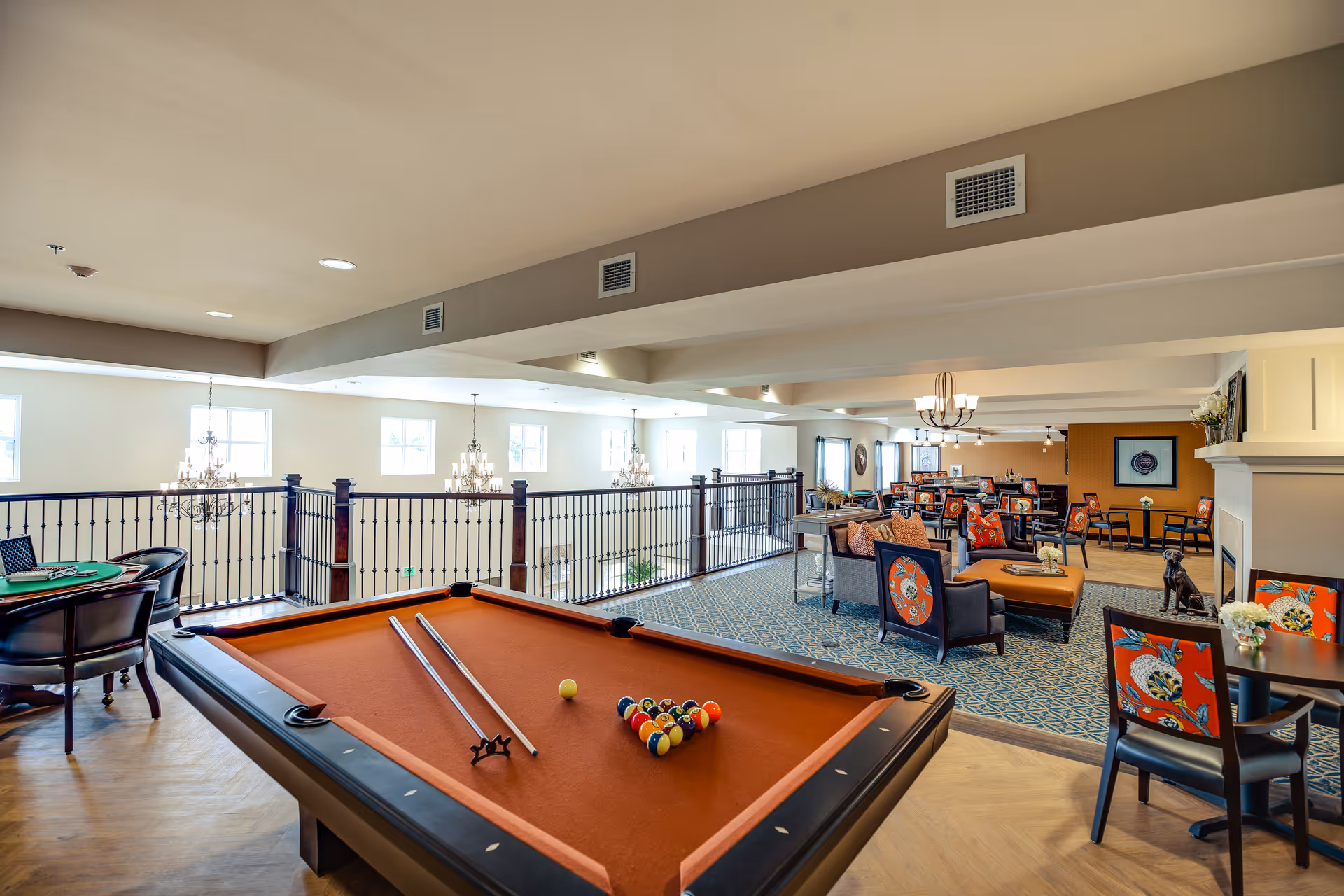 Spacious senior living common area with a pool table in the foreground, seating groups, chandeliers, and a railing overlooking the lower level.