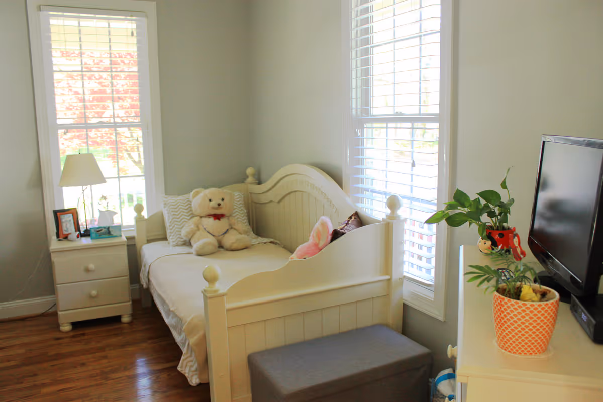 White bedroom with a teddy bear on the bed beside a nightstand and lamp.