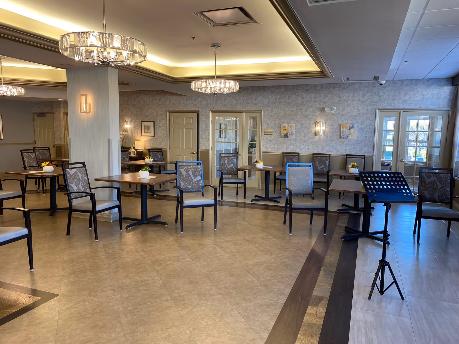 A spacious dining room in an assisted living facility with multiple tables and chairs arranged neatly. The room features modern chandeliers, light-colored walls with framed artwork, and large windows with glass doors letting in natural light. A music stand is positioned near the right side of the room.