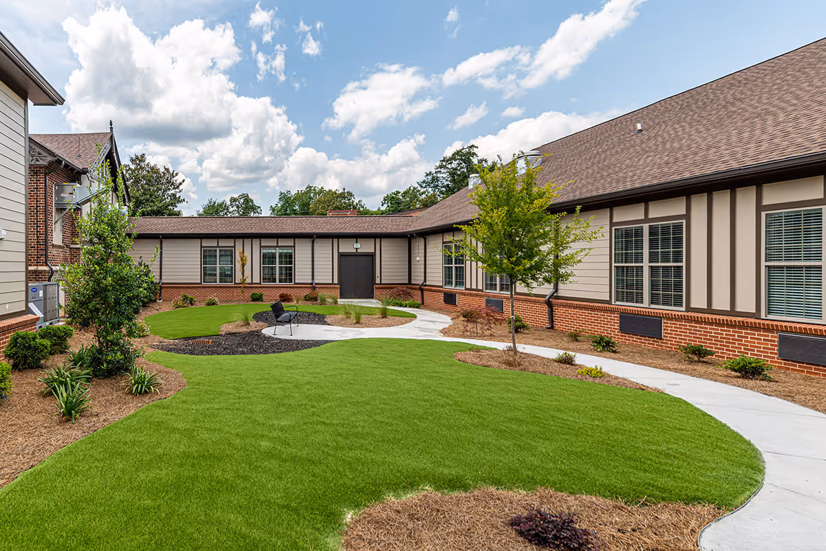 Outdoor courtyard area of Alexander IV facility featuring a well-maintained green lawn, a curved concrete walkway, small trees, shrubs, and a single black chair near the center. The building surrounding the courtyard has beige siding with brick accents and multiple windows under a brown shingled roof. The sky is partly cloudy.
