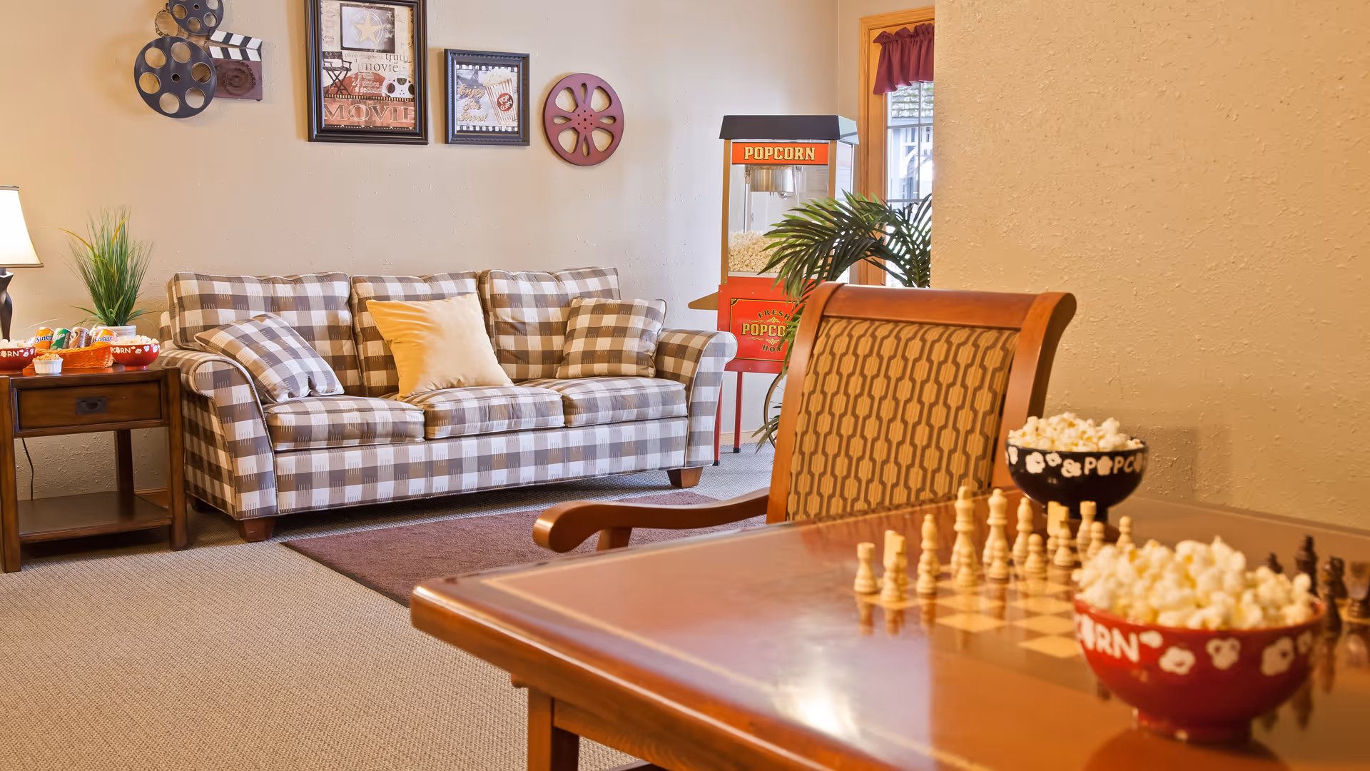 A cozy living room area with a checkered sofa adorned with pillows, a wooden side table with a lamp and snacks, a popcorn machine in the background near a window, and a wooden table with a chessboard and bowls of popcorn in the foreground.