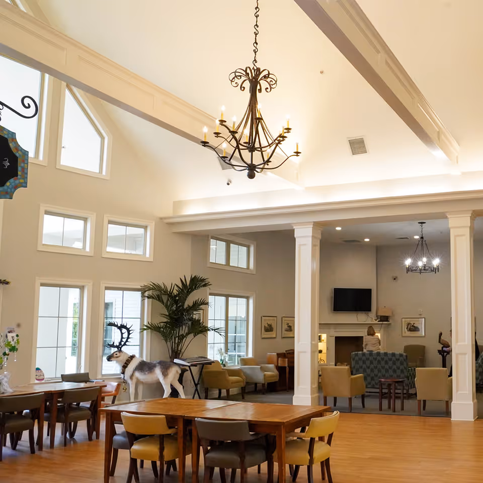 Spacious senior living facility common area with high vaulted ceilings, a large chandelier, wooden tables and chairs, a decorative reindeer statue, potted plants, and a seating area with armchairs and a fireplace in the background.