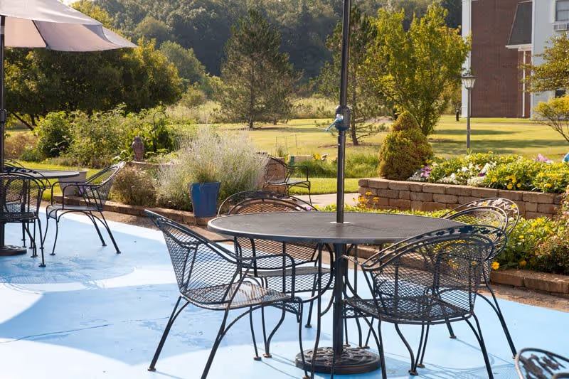 Outdoor patio area with black metal tables and chairs on a light blue concrete surface, surrounded by greenery, plants, and trees with a building partially visible in the background.