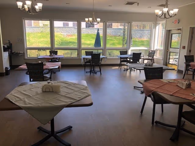A dining room with several tables covered with tablecloths and chairs arranged around them. Large windows along one wall let in natural light and show a grassy outdoor area. Ceiling lights hang above the tables, and there is a door to the right side of the room.
