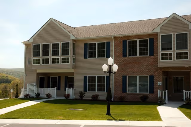 Exterior view of a two-story residential building with beige siding and brick accents, featuring multiple windows with dark shutters, small porches with white railings, a green lawn, and a black street lamp in front.