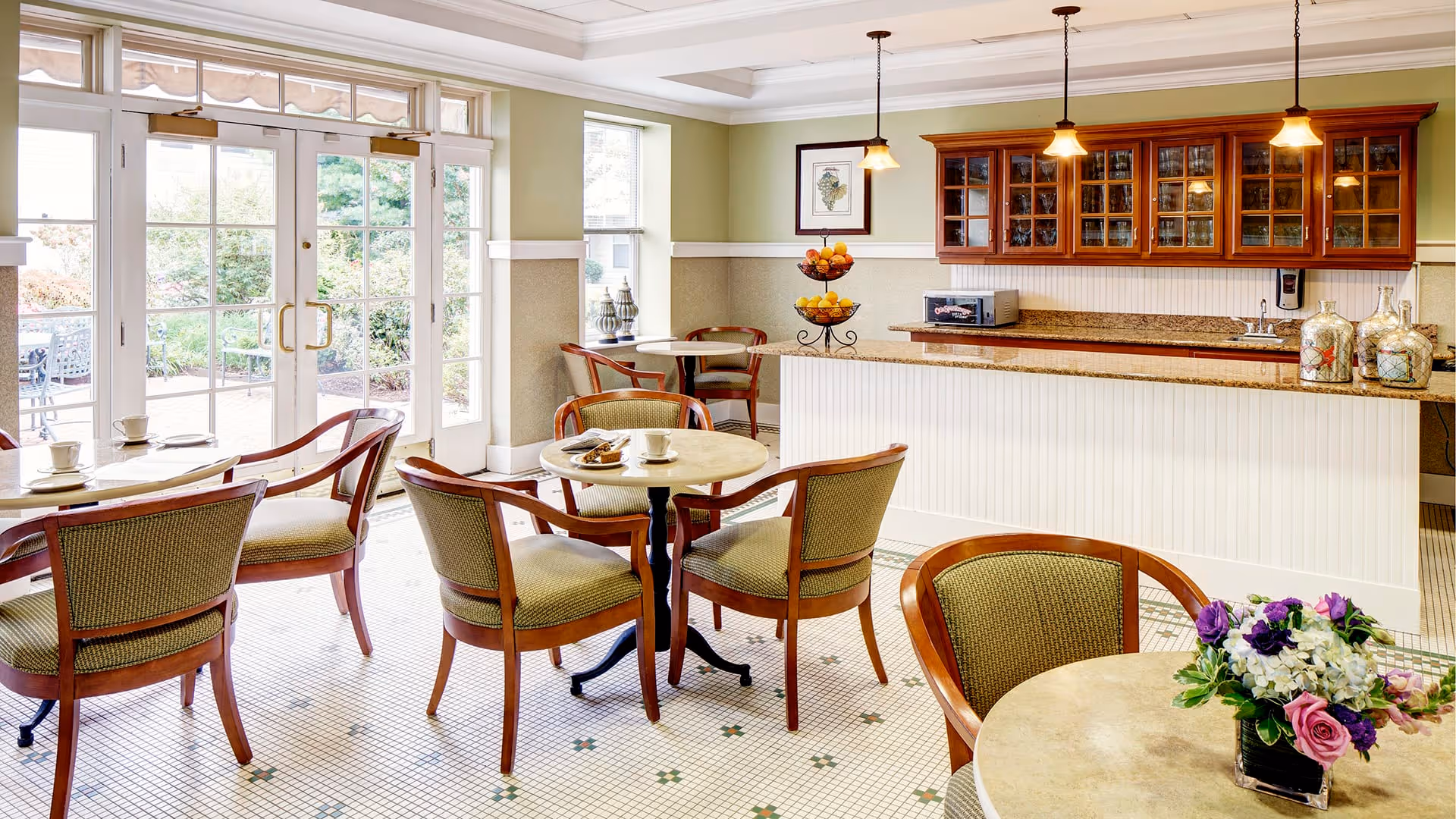 A bright and inviting dining area with several round tables and wooden chairs with green upholstery. There is a counter with a granite top and wooden cabinets above it, along with a microwave and decorative jars. Large glass doors and windows let in natural light and provide a view of an outdoor patio area with greenery.