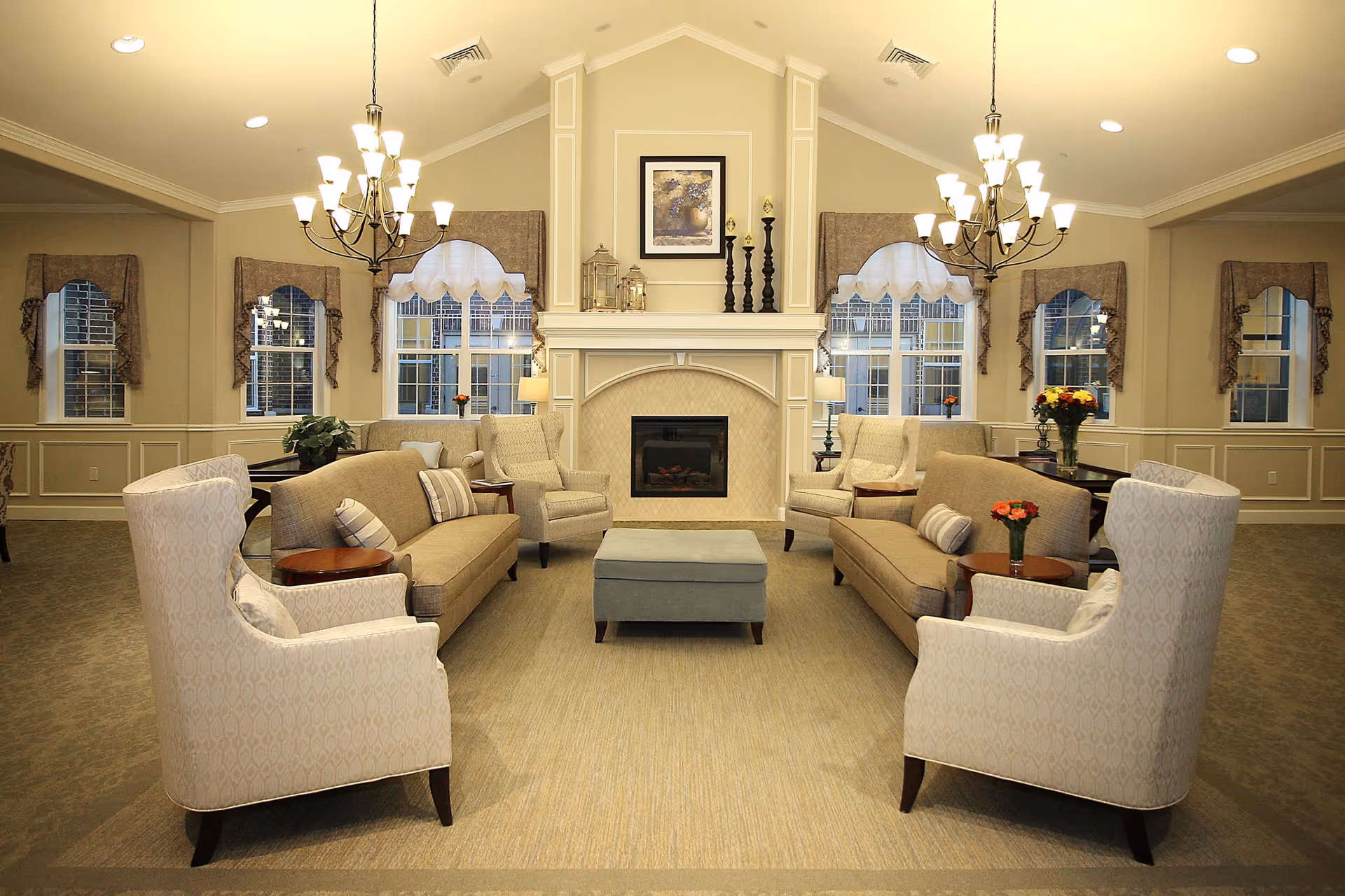 A spacious and elegant senior living common area with two beige sofas, four armchairs, a central ottoman, and side tables with flower vases. The room features a fireplace with decorative items on the mantel, multiple windows with valances, and two large chandeliers hanging from a high ceiling.