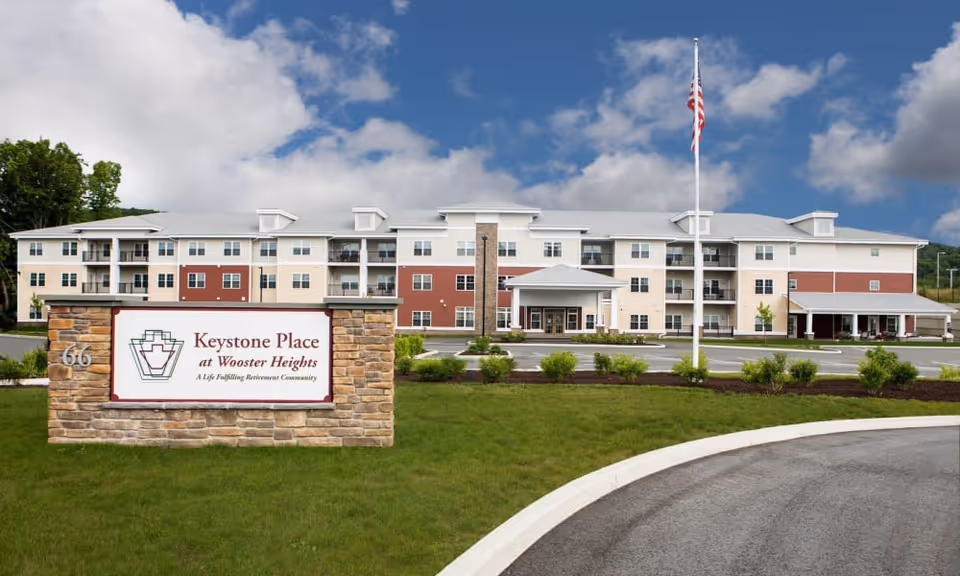 Exterior view of Keystone Place at Wooster Heights, a multi-story retirement community building with a stone sign in front displaying the facility name and logo, surrounded by green grass and landscaping under a partly cloudy sky.