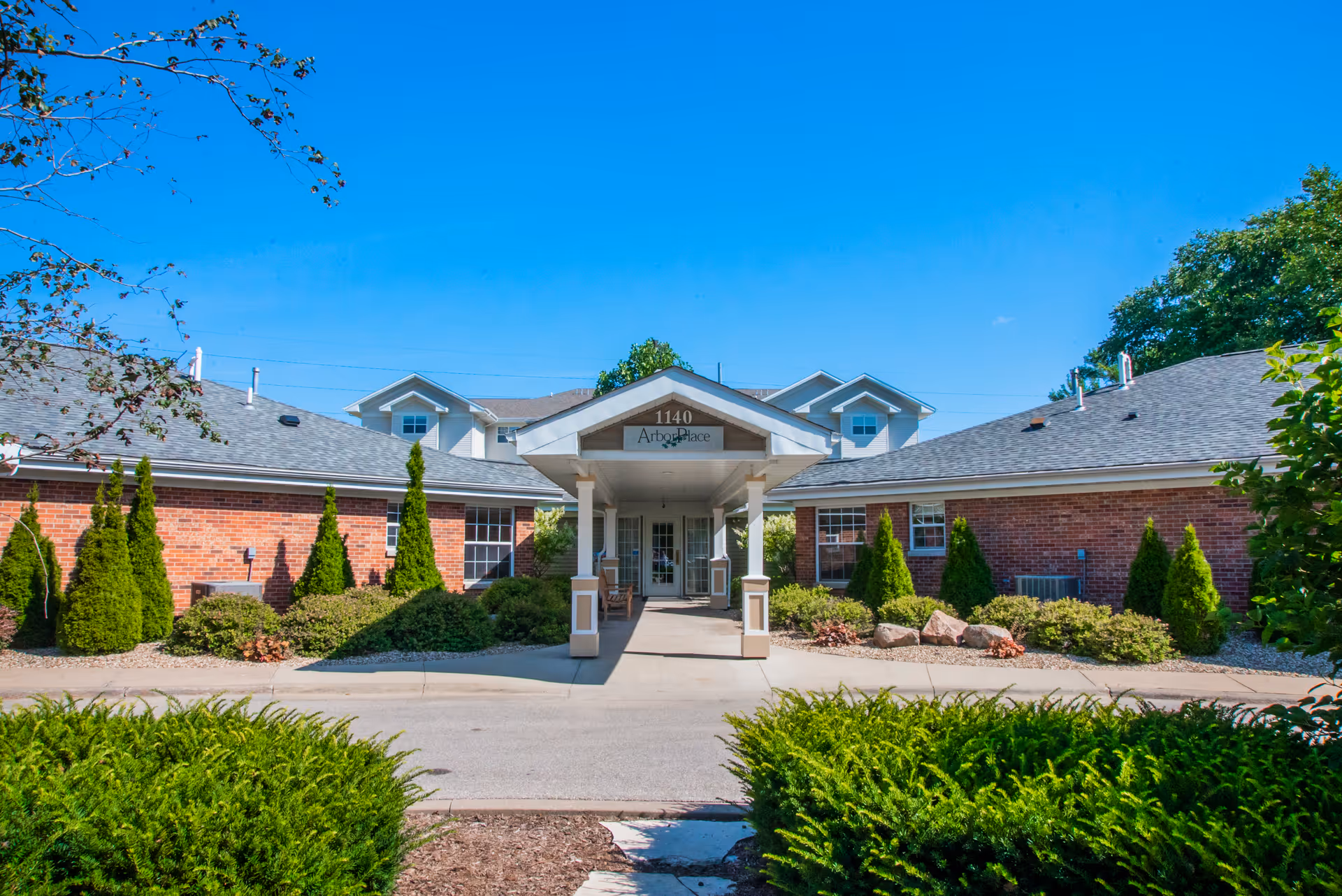 Front exterior view of a single-story brick building with a covered entrance and well-maintained landscaping including bushes and small trees under a clear blue sky.