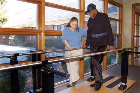 A caregiver assists an elderly man as he walks between parallel bars inside a room with large windows overlooking an outdoor area. The caregiver is supporting the man by holding his arm and guiding him during the walking exercise.
