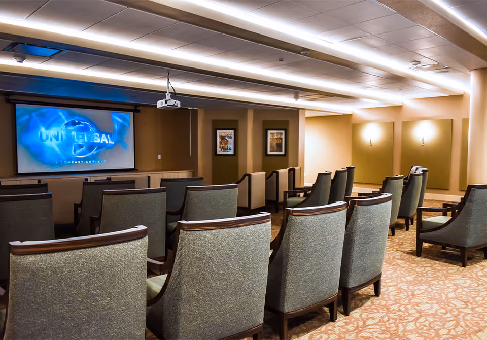 A small theater room with rows of upholstered chairs facing a large screen displaying the Universal logo. The room has warm lighting, framed movie posters on the walls, and a ceiling-mounted projector.