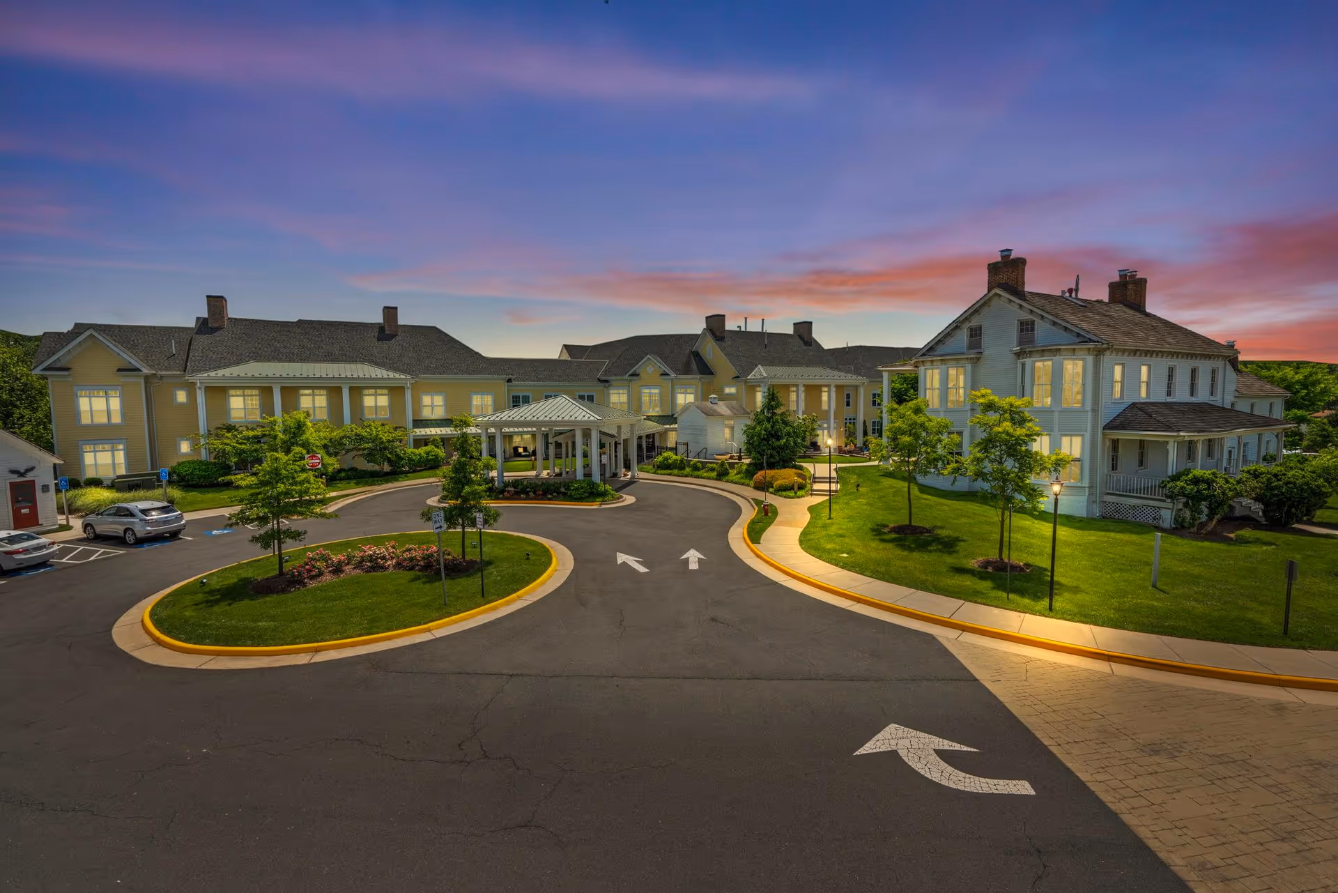 Exterior view of Sunrise at Silas Burke House senior living facility at dusk, showing a large driveway with a roundabout, landscaped greenery, and multiple connected buildings with lit windows under a colorful sky.