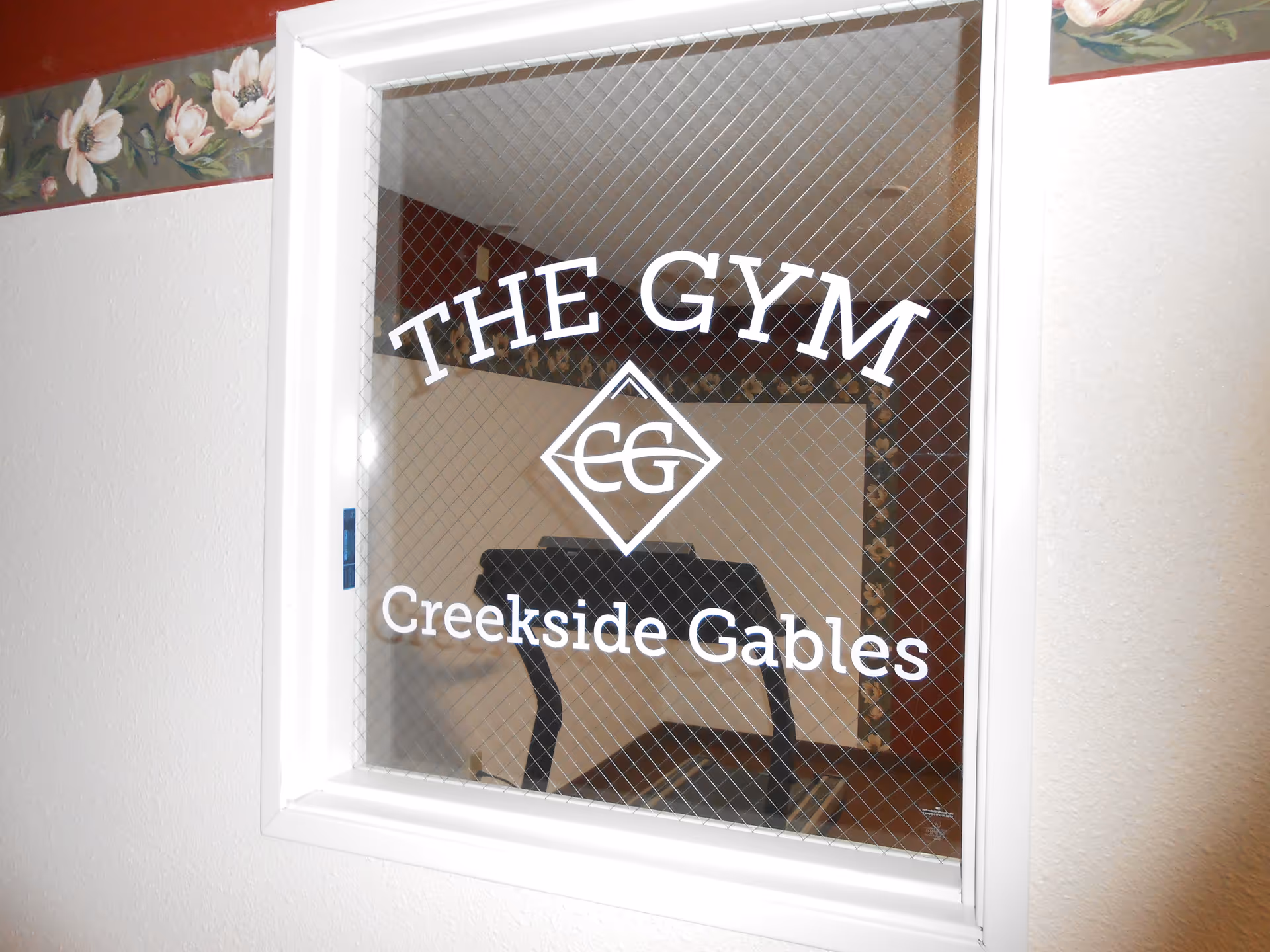 A window with wire mesh showing the words 'THE GYM Creekside Gables' with a logo in the center. Behind the window, a treadmill is visible inside the gym room.