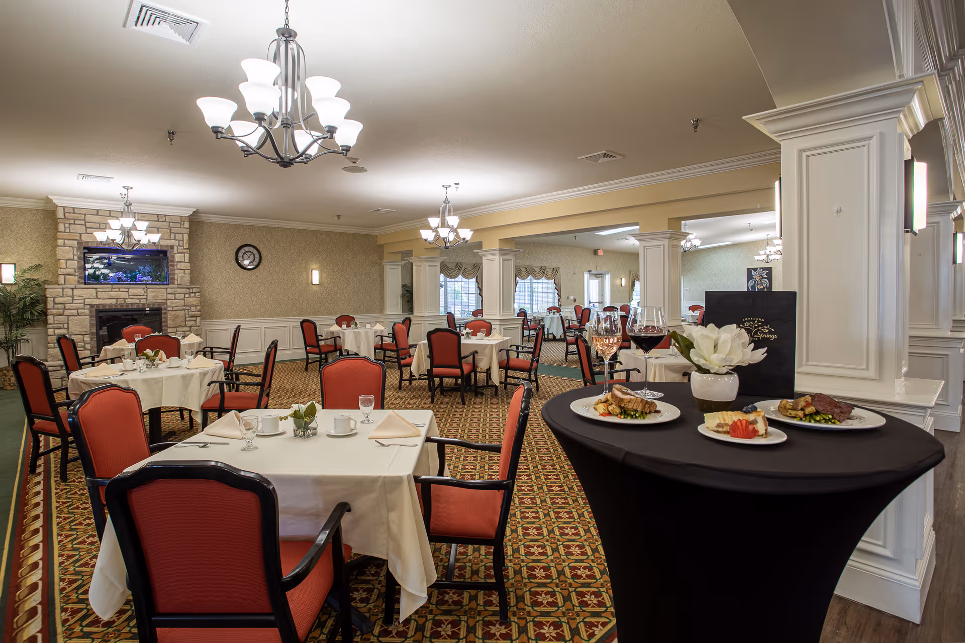 A spacious dining room with multiple round tables covered with white tablecloths and set with cups, glasses, and napkins. The chairs have red upholstery and black frames. There are chandeliers hanging from the ceiling, a stone fireplace with a fish tank above it, and a patterned carpet. In the foreground, a black round table displays three plates of food, two glasses of wine, a white flower in a vase, and a menu.