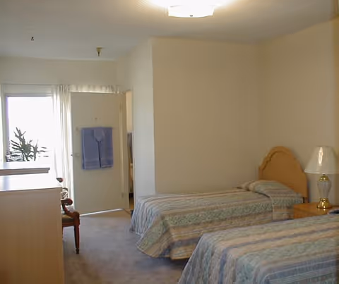 A simple bedroom with two twin beds covered in patterned bedspreads. There is a wooden nightstand with a lamp between the beds. A wooden dresser is on the left side of the room, and a chair is partially visible near a door that leads to another room. A towel hangs on the door, and a window with sheer curtains lets in natural light.