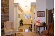 Interior view of a senior living facility entrance area with a front door featuring glass panels. The space includes a small table with a red lamp and framed artwork on the wall. There are two upholstered chairs, one beige and one brown, and decorative columns framing the hallway leading to the door. The floor is hardwood with a rug near the entrance.