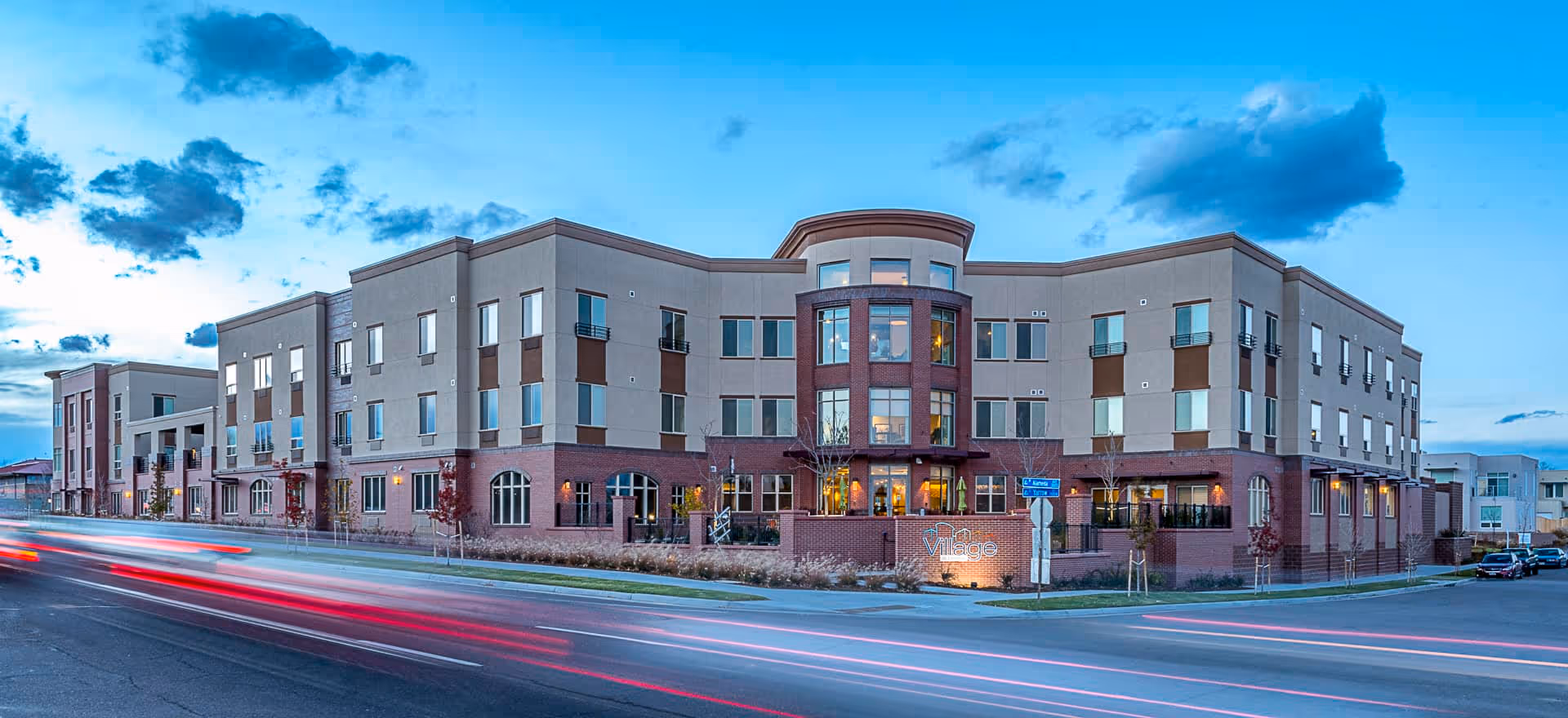 Exterior view of a modern three-story senior living facility building named Village at Belmar during dusk with light trails from passing cars on the street in front.