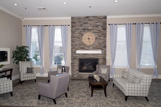 A cozy senior living facility common area with a stone fireplace centered on the back wall, flanked by large windows with light curtains. The room features patterned armchairs and a sofa arranged around a dark wooden coffee table on a patterned carpet. A large round clock is mounted above the fireplace, and a potted plant is visible in the corner.