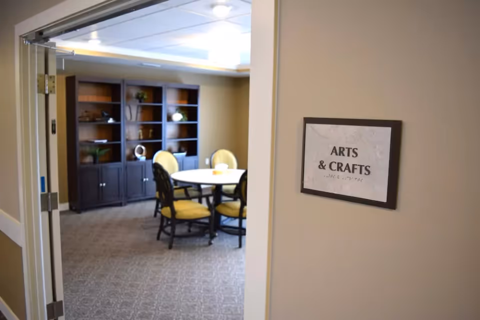 View through an open doorway into an arts and crafts room with a round table surrounded by four yellow cushioned chairs and a dark wooden bookshelf against the far wall.