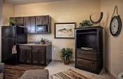 Interior view of a senior living facility room featuring a kitchenette with dark wooden cabinets, a black refrigerator, microwave, and sink. There is a framed fish artwork on the wall above a small plant, a dark wooden TV stand with a flat-screen TV, a wall clock, and a patterned rug on the floor.