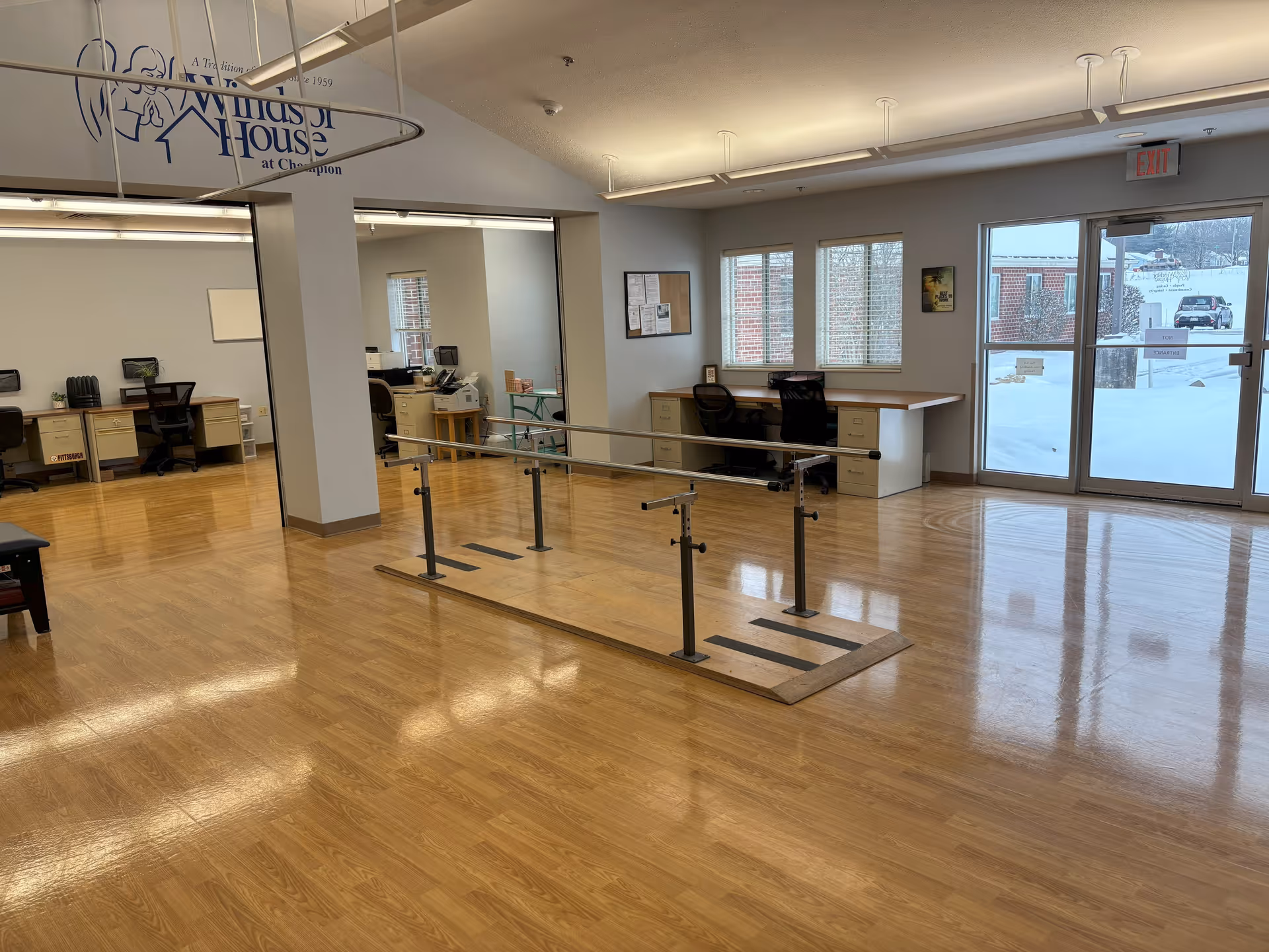 A spacious room with polished wooden floors and a set of parallel bars in the center, likely used for physical therapy or rehabilitation. The room has several desks and office chairs along the walls, large windows letting in natural light, and a glass door showing a snowy outdoor scene. The ceiling has fluorescent lighting, and there is a Windsor House at Champion logo visible on the upper left side of the image.