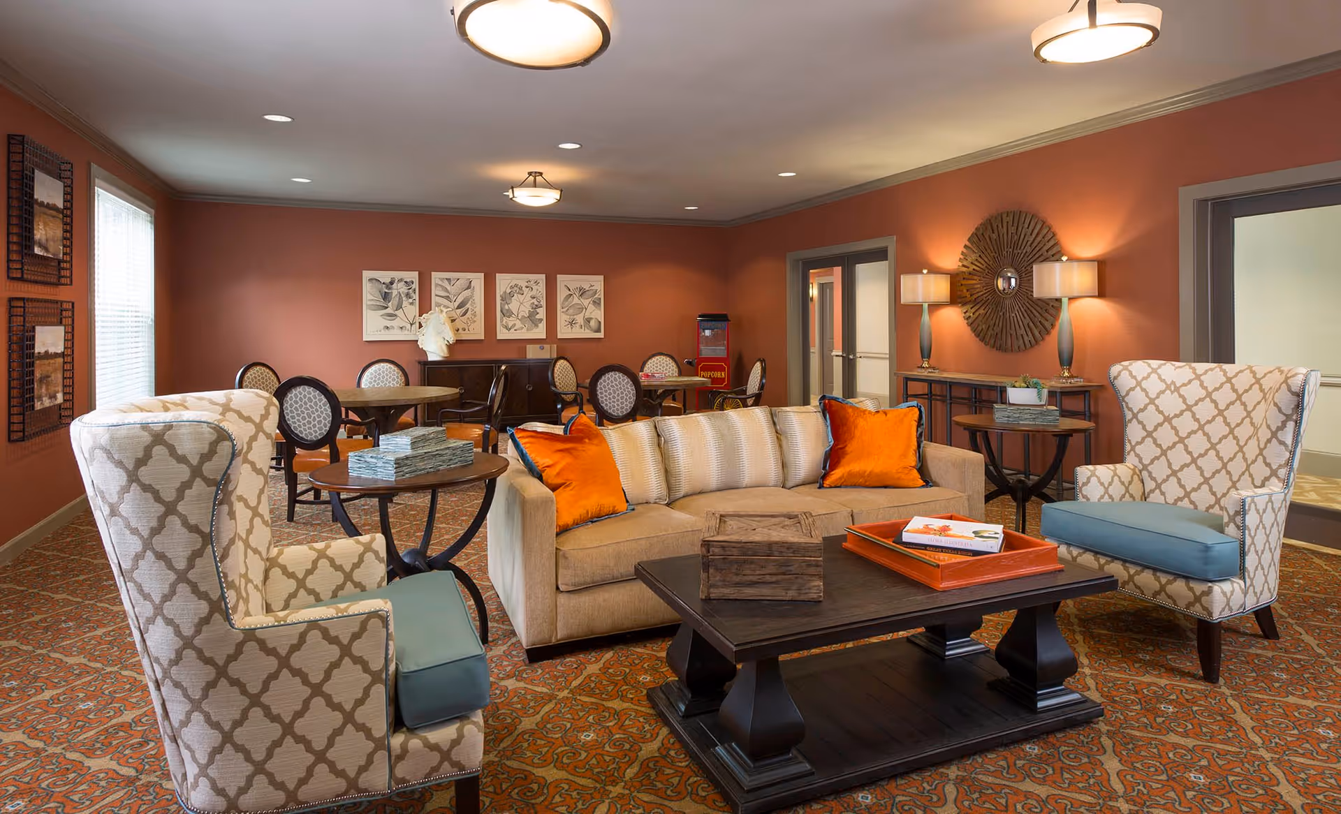 A cozy living room area in an assisted living facility with a beige sofa adorned with orange and beige pillows, two patterned armchairs with blue cushions, a dark wooden coffee table with books and a tray, and a side table with stacked books. The room has warm terracotta-colored walls, decorative framed artwork, a patterned carpet, and soft lighting from ceiling fixtures and table lamps. In the background, there are round tables with chairs and a popcorn machine near the wall.