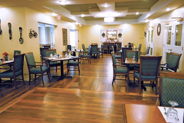 Spacious dining room with multiple set tables and upholstered chairs on a wood floor under recessed ceiling lights.