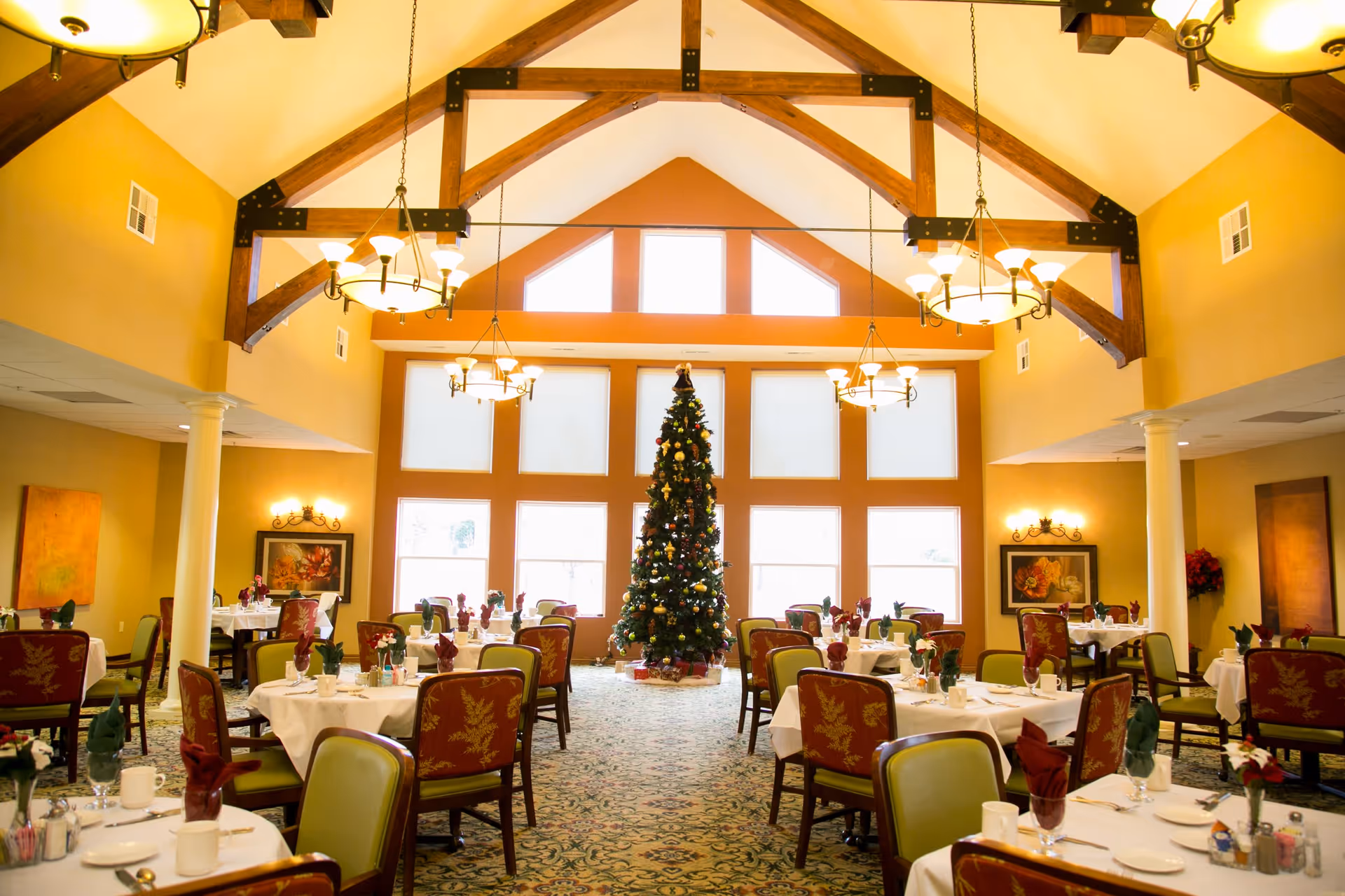 A spacious dining room decorated for the holidays with a tall Christmas tree in the center near large windows. The room features high vaulted ceilings with exposed wooden beams and multiple chandeliers. Tables are set with white tablecloths, napkins, and dining ware, surrounded by chairs with red and green upholstery. Warm yellow walls and framed artwork complete the cozy atmosphere.