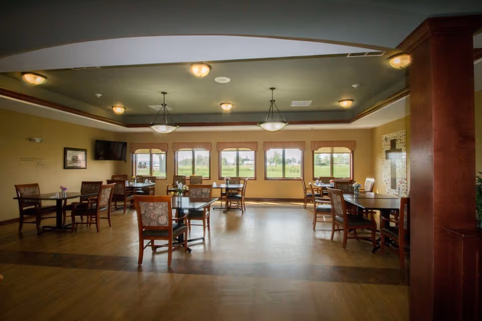A spacious dining room with several wooden tables and chairs arranged neatly. The room has large windows with a view of green outdoor space, allowing natural light to fill the room. The ceiling features multiple light fixtures, and there is a wall-mounted TV on one side. The floor is wooden, and the walls are painted in warm tones.