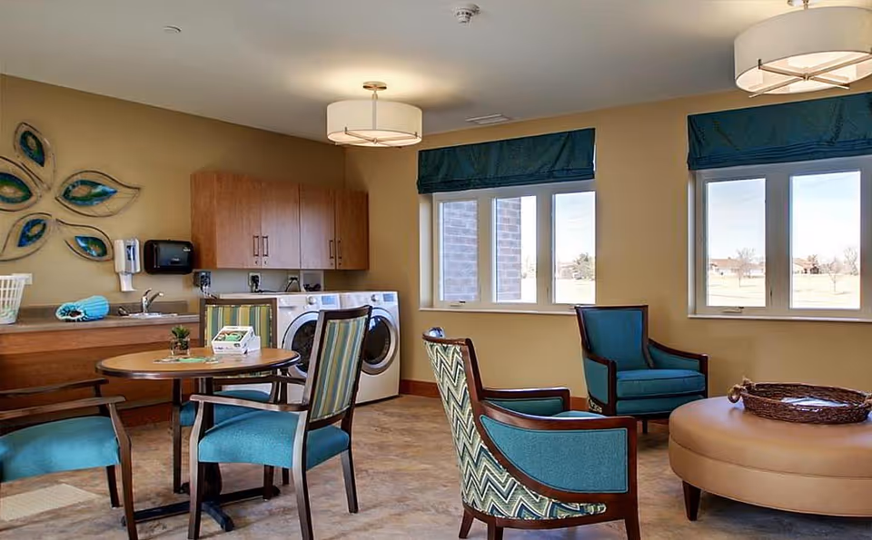 A bright and cozy room in Evergreen Senior Living featuring a small round table with four chairs, two teal armchairs, a large round ottoman with a wicker tray, a laundry area with a washer and dryer, wooden cabinets, and two windows with blue valances letting in natural light.